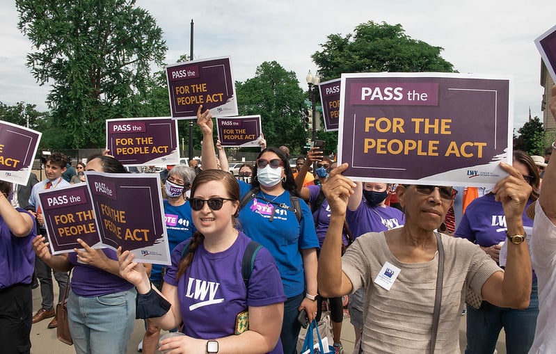Women stand at a rally holding signs promoting the For the People Act
