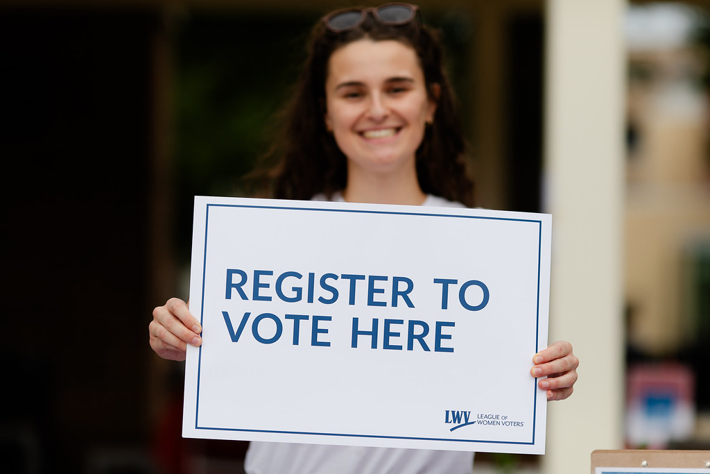 A woman holding an LWV sign that says "Register to Vote Here"