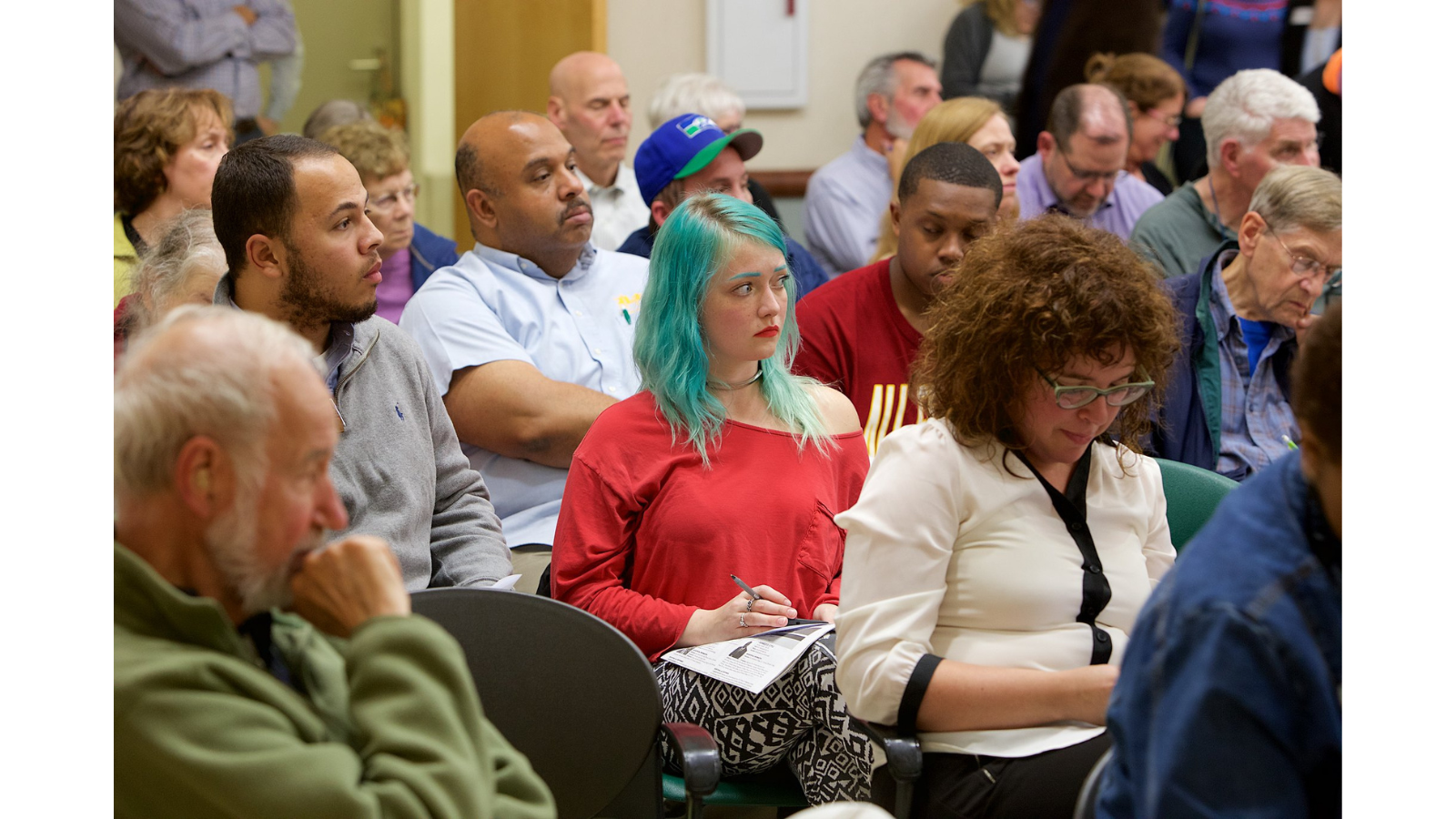 A woman with blue hair is sitting in the audience of a candidate forum.