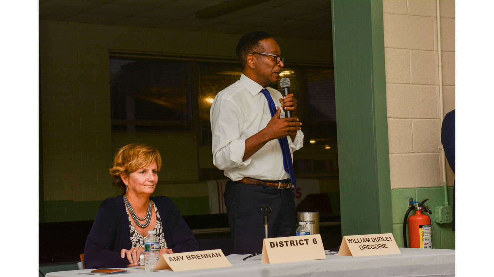 Two candidates are behind a desk at a candidate forum. On the left is a woman, sitting down. She has a water bottle and candidate name tent in front of her, the name tent says "AMY BRENNAN". On the right is a man standing up, speaking into a micrphone. He has two name tents in front of him on the table: one says "DISTRICT 6" and another says "WILLIAM DUDLEY GREGORIE."