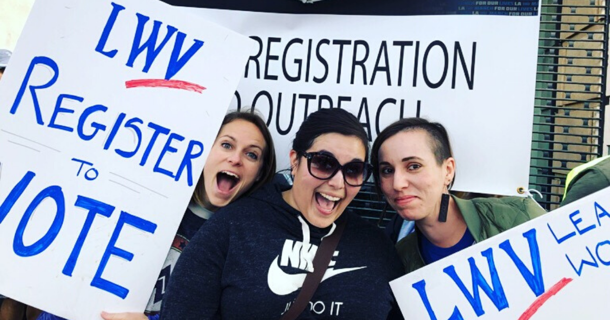Three young women pose together outdoors, smiling and excited, and holding hand-made signs with the League of Women Voters logo that say "Register to Vote"