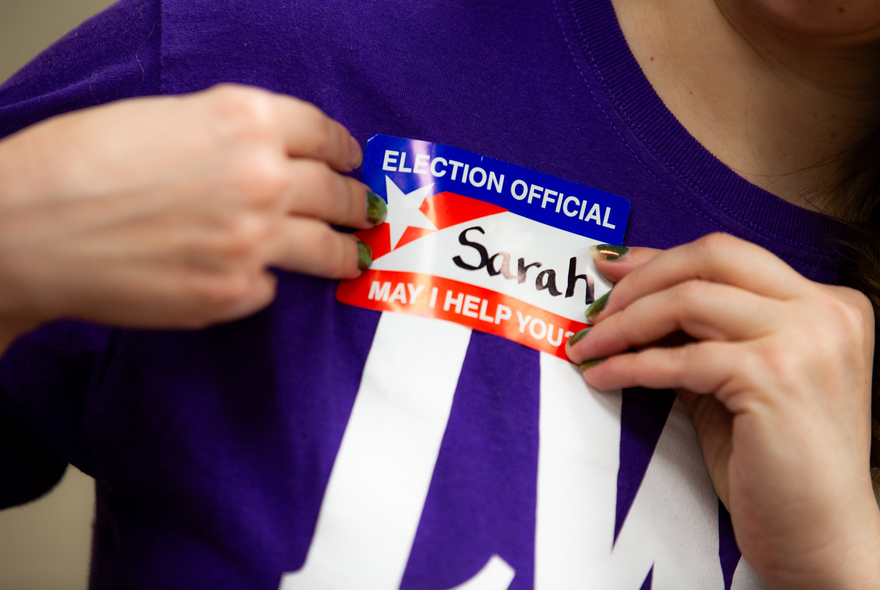 A person in a purple LWV short putting on an election official badge that says "My name is Sarah"