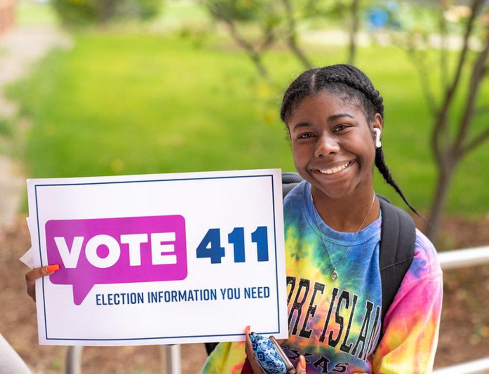 Girl smiling and sitting next to a sign that says "VOTE411"