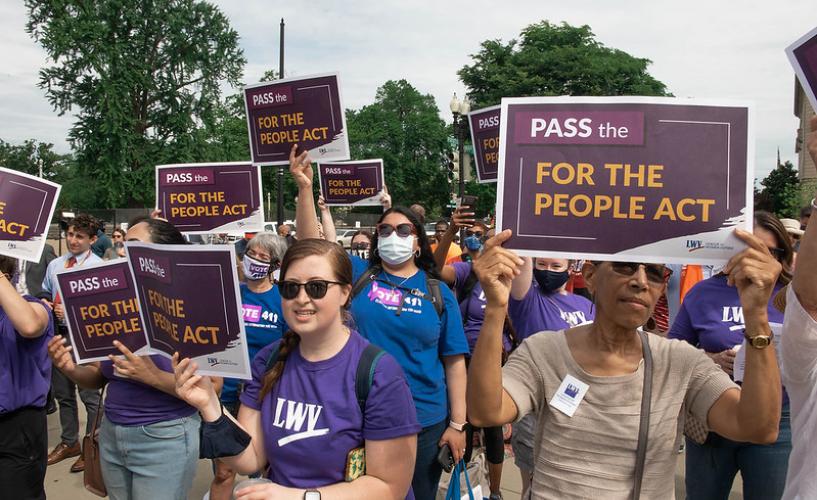 Women stand at a rally holding signs promoting the For the People Act
