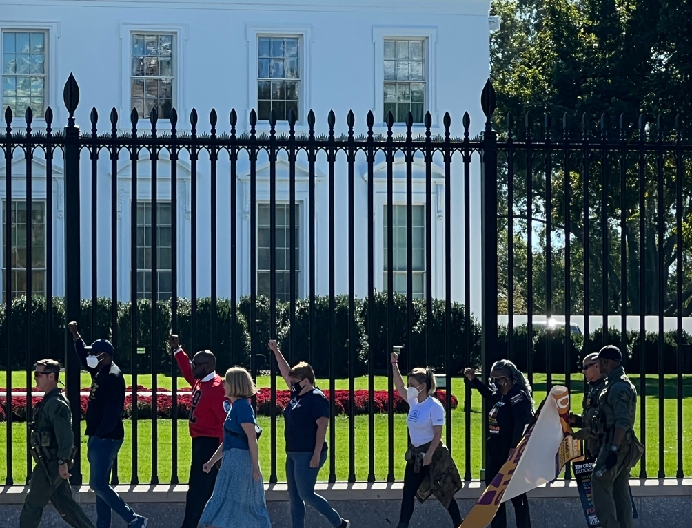 Protestors walking in a line in front of the White House with their fists up; Polic officer ushering them forward for arrest
