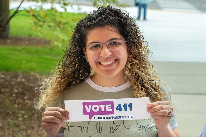 Woman holding up a bumper sticker that says "VOTE411"