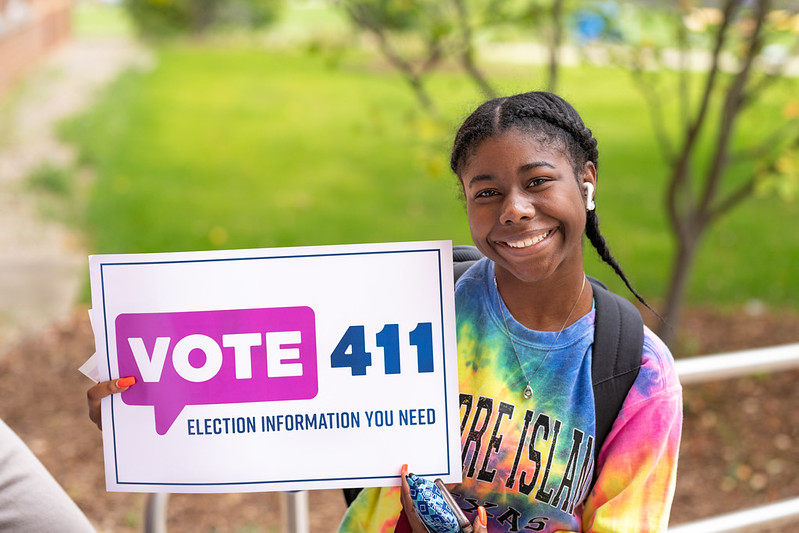 Girl smiling and sitting next to a sign that says "VOTE411"