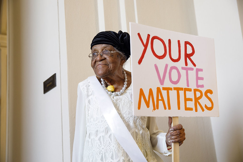 An elderly woman holding a sign that says "Your Vote Matters"