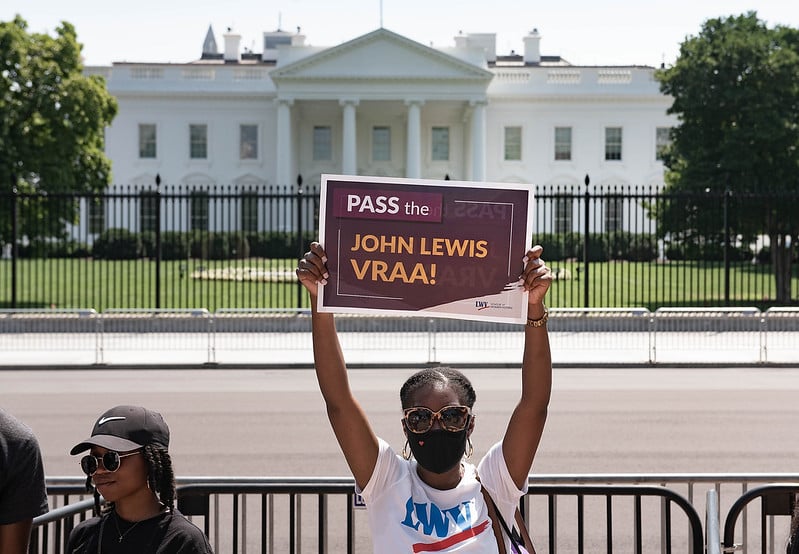 A woman in a LWV T-shirt in front of the White House holding a sign that says "Pass the John Lewis VRAA"