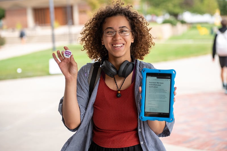 A young woman holding a VOTE411 sticker and an ipad showing she registered to vote