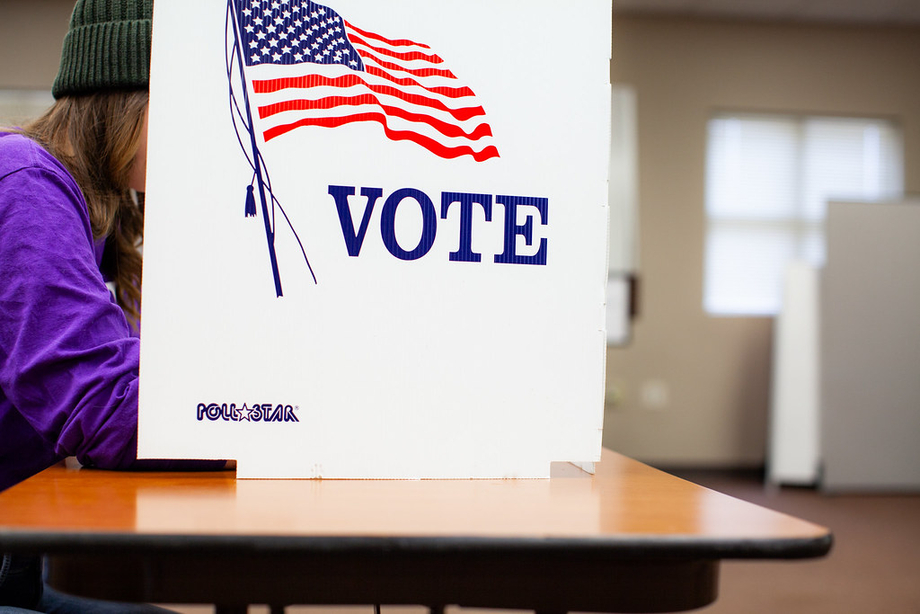 A person is voting behind a white sign with an American flag that says "VOTE"