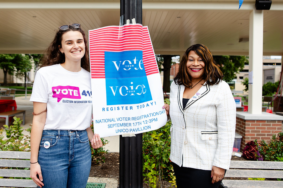 Two women, one wearing a VOTE411 shirt, standing next to a sign promoting voter registration
