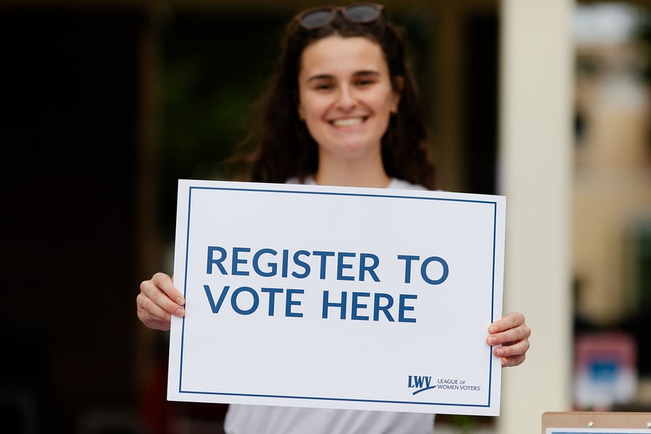 A woman holding an LWV sign that says "Register to Vote Here"