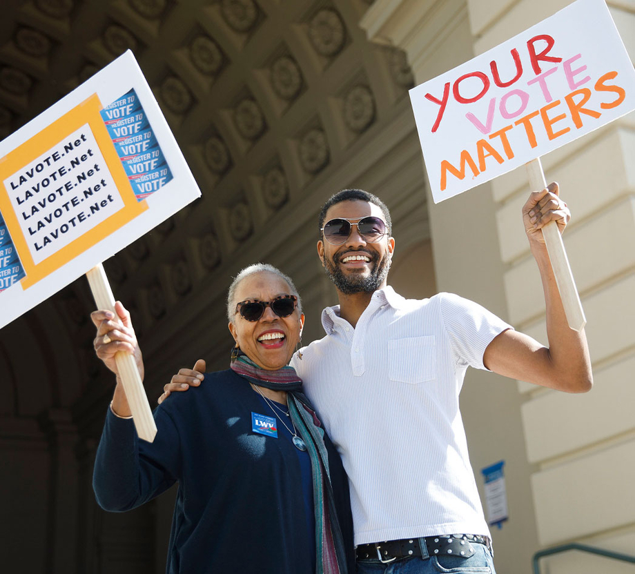 An older woman holding a sign, side hugging a young man also holding a sign that reads "your vote matters" outside