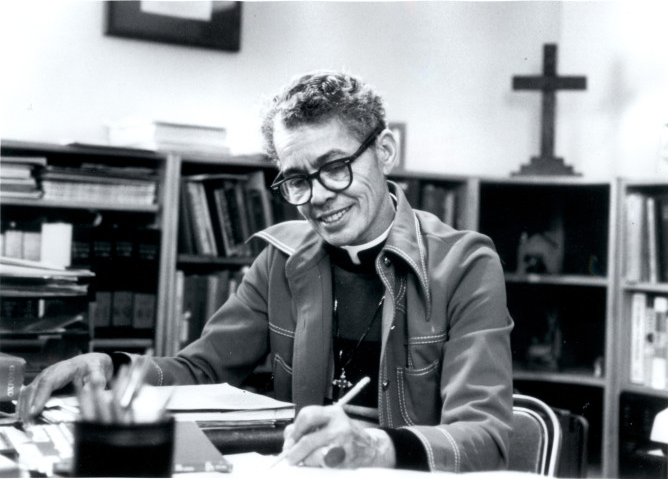 Pauli Murray writing at a desk with a cross on a table behind her