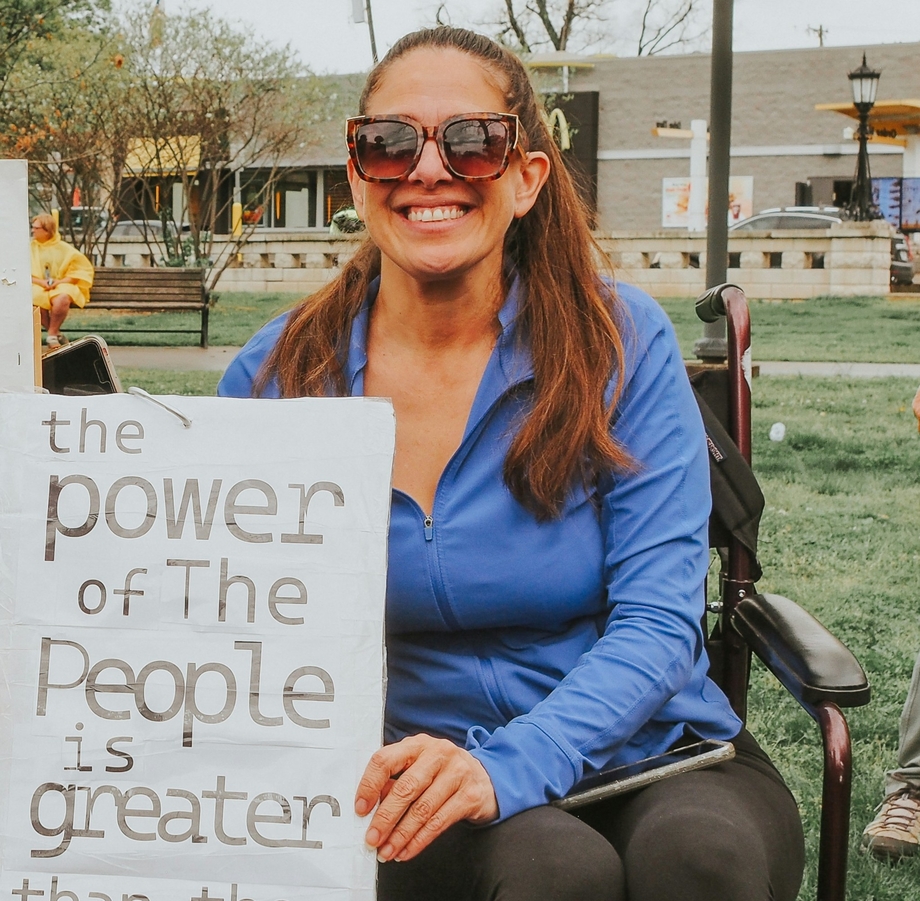 A smiling woman in a wheelchair holding a sign that says "the power of the people is greater than the people in power"