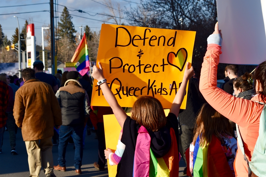 Protestors, one of whom holds a sign saying "Defend + Protect Queer Kids"
