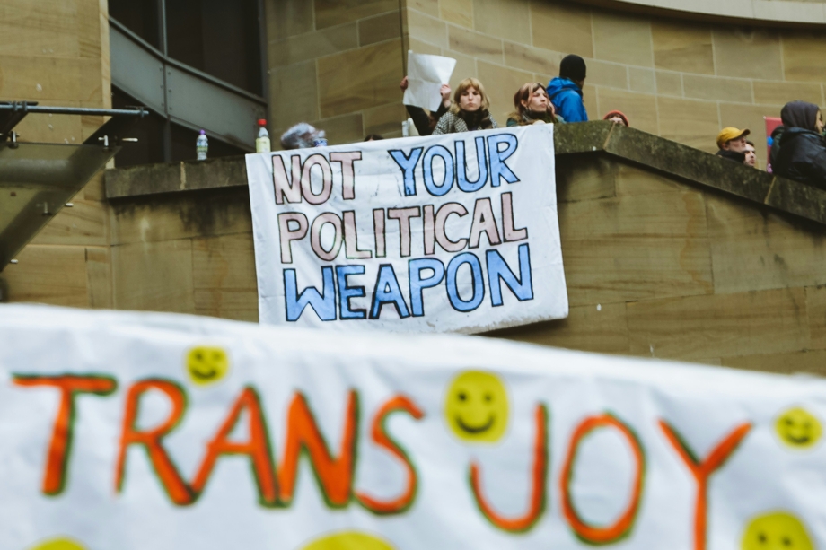 Protestors above a banner that says "Trans Joy" holding a sign that says "not your political weapon"