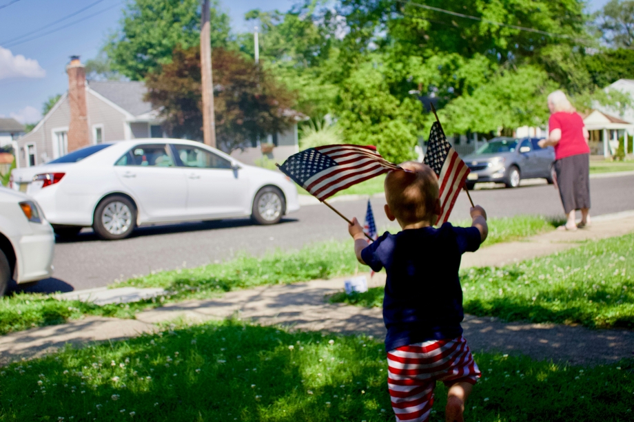 A toddler on a lawn holding up American flags in both hands