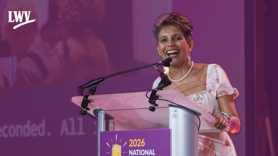 Board President Sania Irwin speaking at a podium on a purple backdrop with the white LWV logo in the top left corner