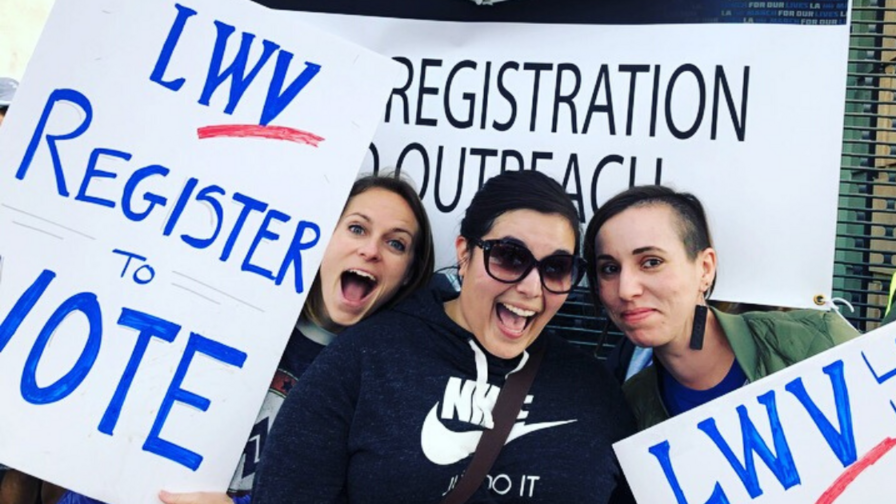 Three young women pose together outdoors, smiling and excited, and holding hand-made signs with the League of Women Voters logo that say "Register to Vote"